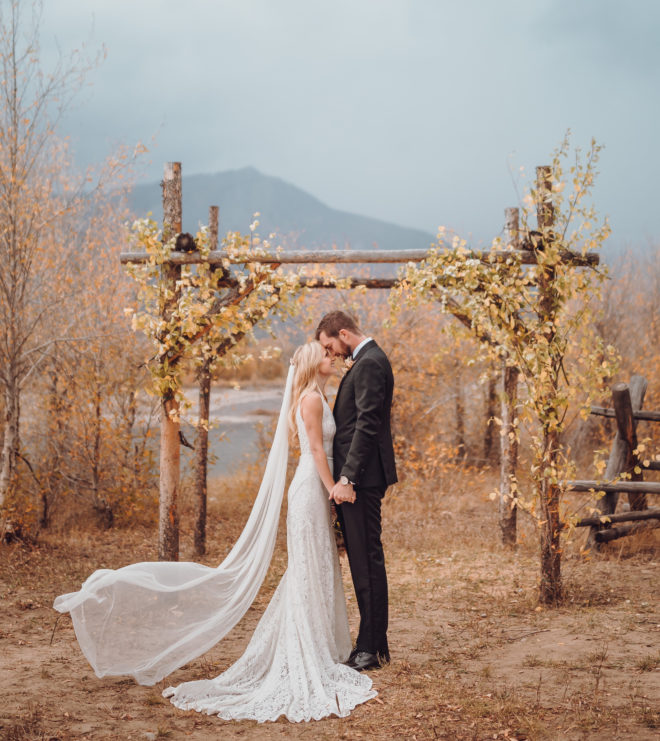 Bride, in long fitted lace white wedding gown and long veil, stands forehead to forehead with a groom, wearing a black suit, in front of an altar created from golden-leafed aspens beside the Snake River in a Jackson Hole wedding ceremony.