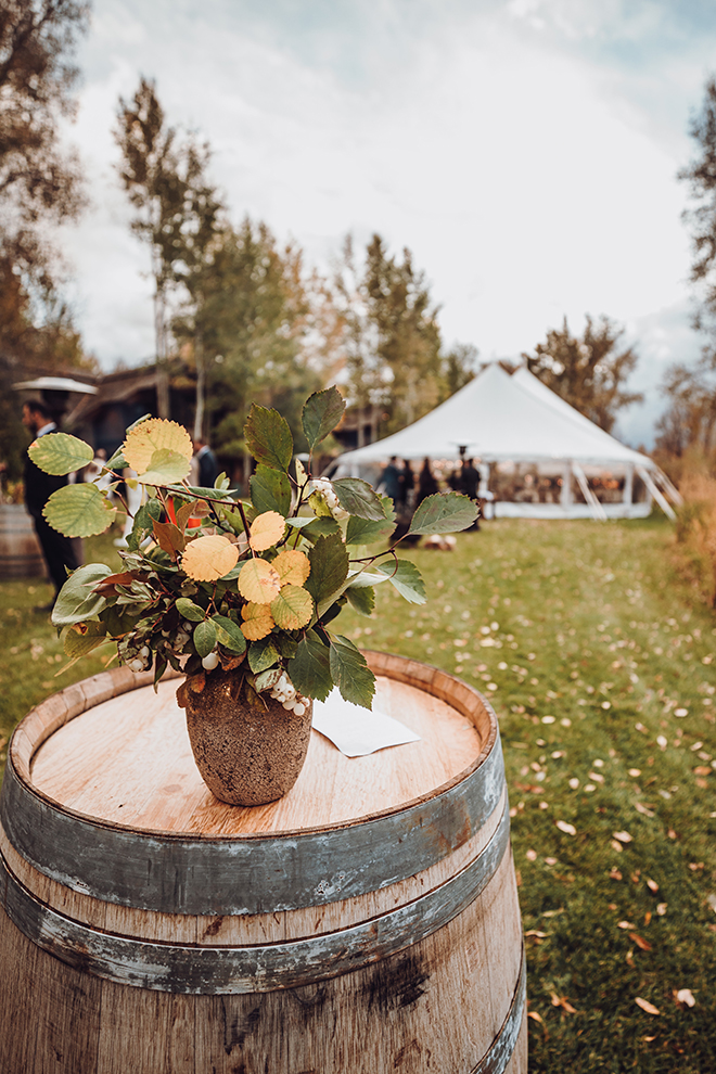 A wine barrel adorned with sprigs of aspen leaves and berries rests atop a wine barrel with a tented reception in the background.