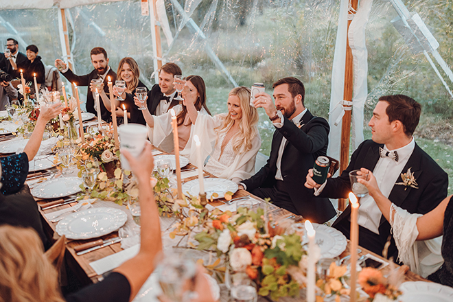 The bride and groom join their friends and family for a toast while seated in the center of a long reception table adorned with fall blooms and aspen sprigs.