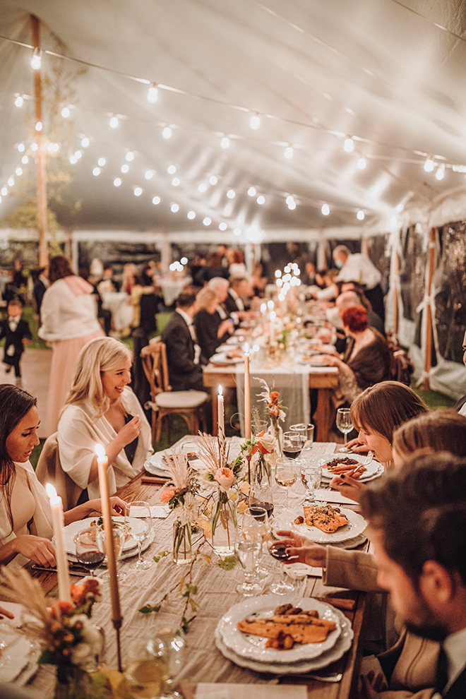 Seated guests at a long reception table adorned with lit tapered candles and florals underneath string lights inside a tent at a Jackson Wyoming wedding. 