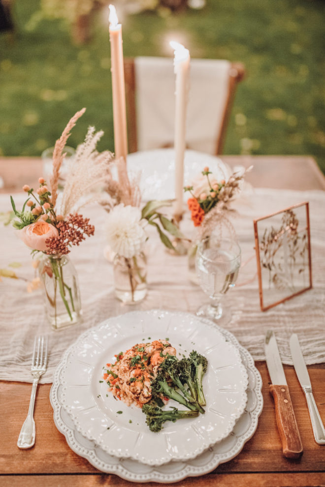 Place setting with white scalloped plate and charger topped with a green vegetable and a serving of meat on a wooden table set with small bud vases filled with pink, mauve and cream colored botanicals, two lit tapered candles and a frame showcasing pressed florals at a Jackson Hole, Wyoming wedding. 