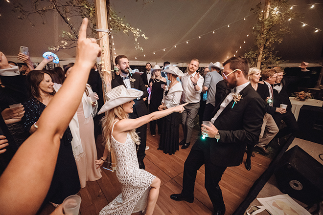 A bride wearing a sequin cowboy hat and dress dances and points at a groom wearing glasses made of glow sticks inside a reception tent during an intimate Jackson Hole Wyoming wedding. 