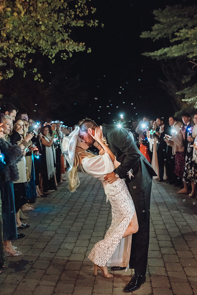 Bride in white fitted gown with deep slit and white glittered cowboy hat kisses groom during a their wedding send-off at their Jackson Wyoming wedding. 