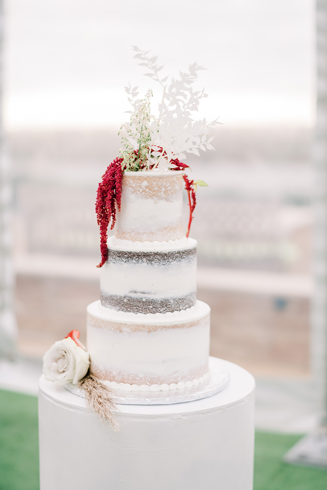 A three-tiered naked wedding cake by Susies Cakes is adorned with a white rose bloom and a blade of pampas grass, including red Amaranthus and white botanicals on the top tier.