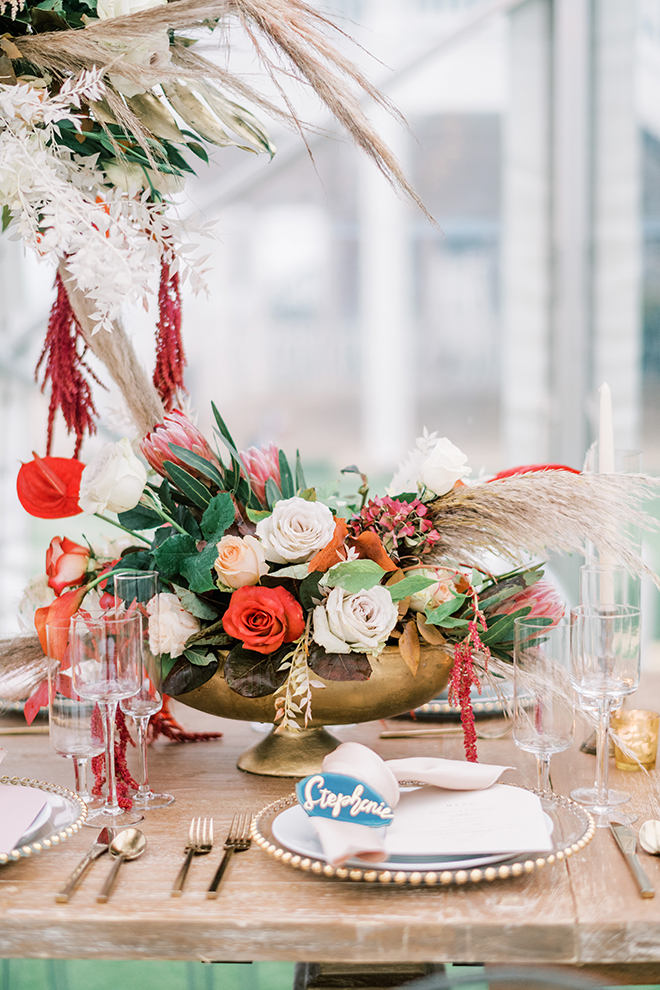 Small organic floral centerpiece in footed gold bowl aside a large pampass grass adorned with red,white and green botanicals atop a washed wood reception table set with gold adorned place settings and tall tapered candles. 