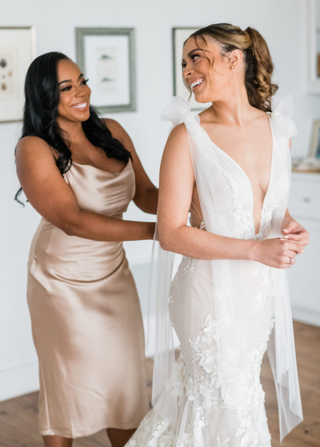 Bridesmaid with long curled hair and short silk blush dress buttoning the brides white Galia Lahav gown with a deep plunging neckline and tied straps, while the bride smiles back.