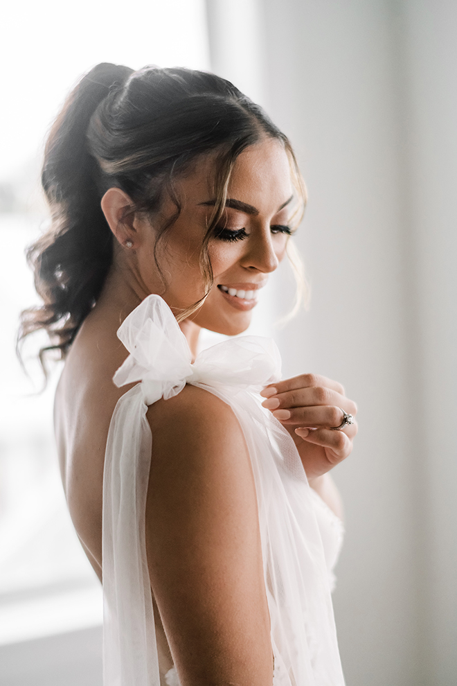 The brides dress has a sweet tulle bow on the right shoulder as natural light casts an ethereal glow over her.