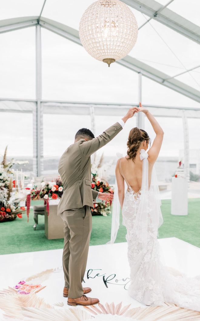 Groom in brown suit twirling a bride with a long deep v-back wedding gown underneath a statement light inside a sun dappled tented wedding space by Galveston Bay.