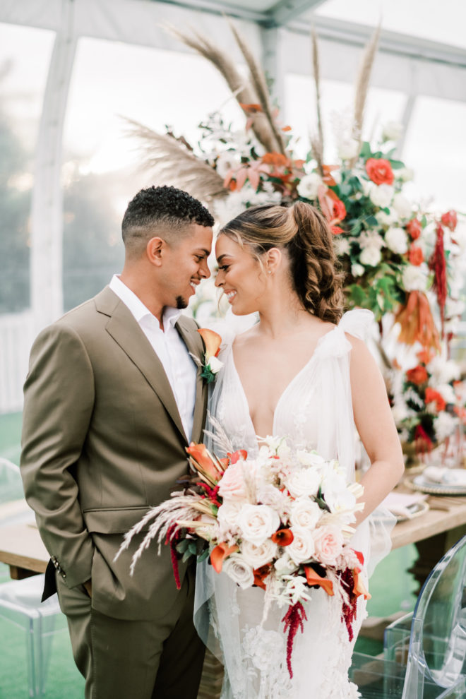 Smiling Groom with his body turned towards a smiling bride holding a lush bouquet of orange, red and blush colored botanicals inside a tented reception space with tall pampas grass and floral centerpieces, long farmhouse tables and ghost chairs on a private property next to Galveston bay in Galveston Texas.