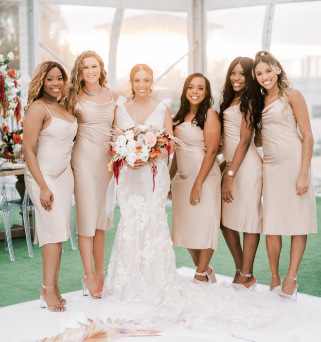 Five bridesmaids wearing fitted blush slip dresses smile with the bride wearing a plunging neckline gown by Galia Lahav while holding a lush bouquet of pampas grass, red botanicals and orange and blush colored florals inside a tented wedding reception designed by Plants N Petals.