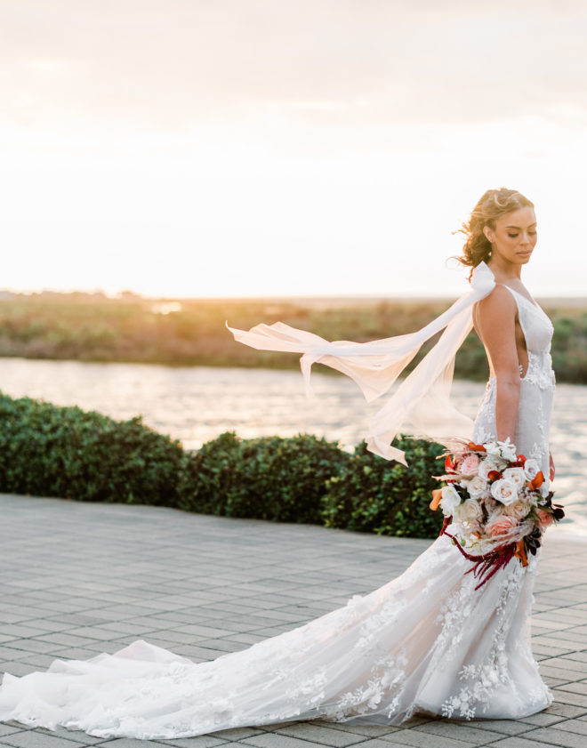 Bride in Galia Lahav fitted gown with long sweeping train and long sleeves blowing in the wind while holding a lush bouquet of sunset-hued florals with the waters of Galveston Bay behind her during the sunset hour.