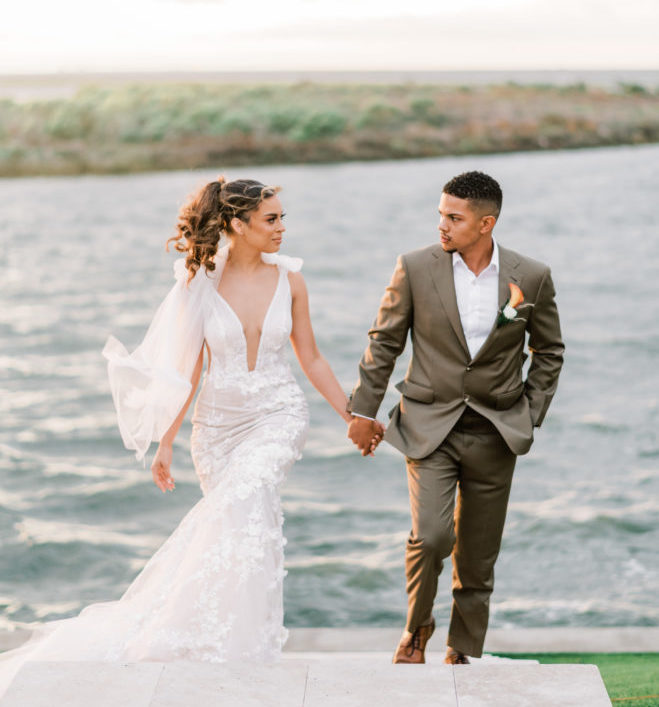 Bride in plunging neckline wedding gown by Galia Lahav holding hands with a groom while walking up a set of outdoor stairs with Galveston Bay behind them.