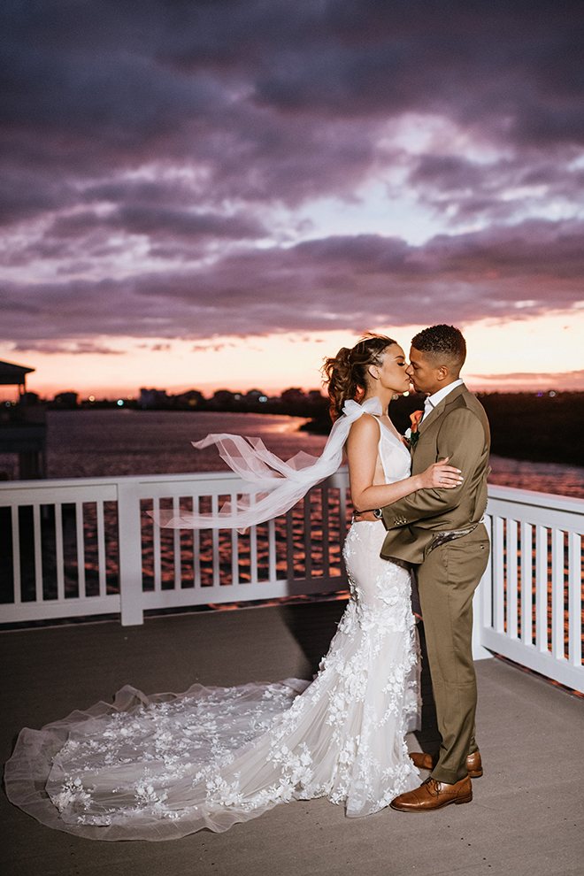 The bride and groom exchange a kiss on the balcony of the Galveston Bay-side home as the sunset paints the sky rich shades of purple, orange, and peach.