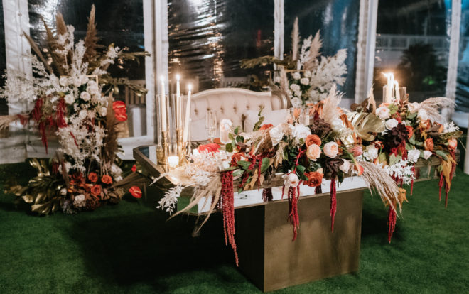 Head table with khaki colored settee set behind a gold mirror top tabled adorned with pampas grass, greenery, orange, white, red and blush roses and lit tapered candles during a tented reception designed by Plants N Petals. 