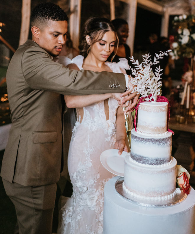 Groom and bride cut into a three tiered lightly iced cake with white and red botanicals atop the first tier by Houston Baker, Susies Cakes.