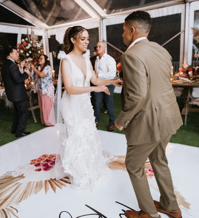 bride in white Galia Lahav dress with tied shoulder straps dances with the groom in a light brown suit on a white dance floor that reads the couples last name, "The Roys" inside their tented reception in Galveston.