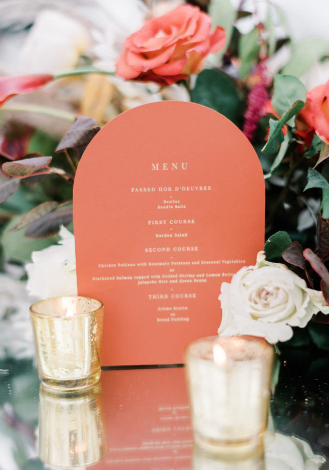 Coral colored half circle menu that reads, "Menu" and "Passed Hor Doeuvres" nestled between coral, blush and orange florals and two gold lit candle votives in an intimate tented ceremony designed by Plants N Petals. 