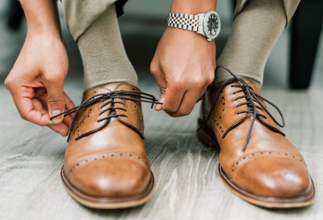 Groom with silver watch on his wrist tying the laces of light brown loafers.