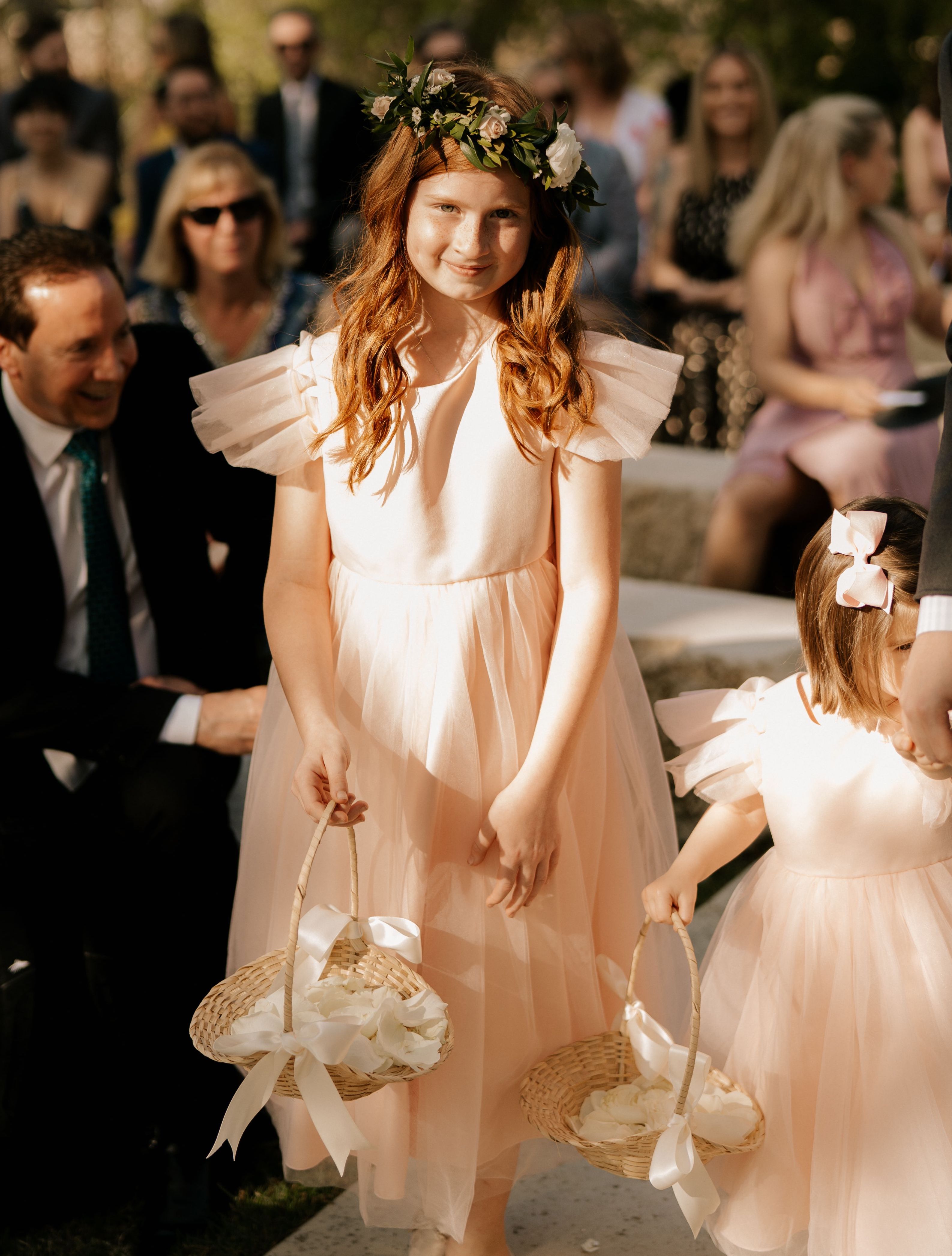 An adorable flower girl walks down the aisle wearing a floral crown and blush flowy dress.