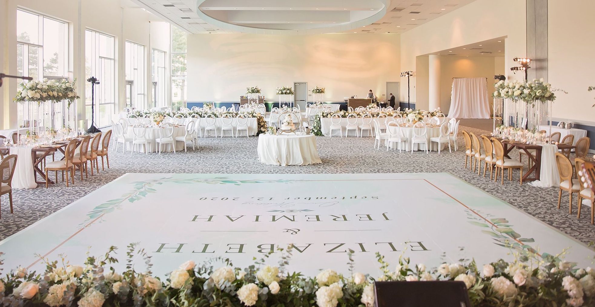 The Del Lago ballroom at the Margaritaville Resort, Lake Conroe is decorated in white rose and eucalyptus floral arrangements, off-white linens and a white dance floor with a personalized decal of the bride and groom's name. 