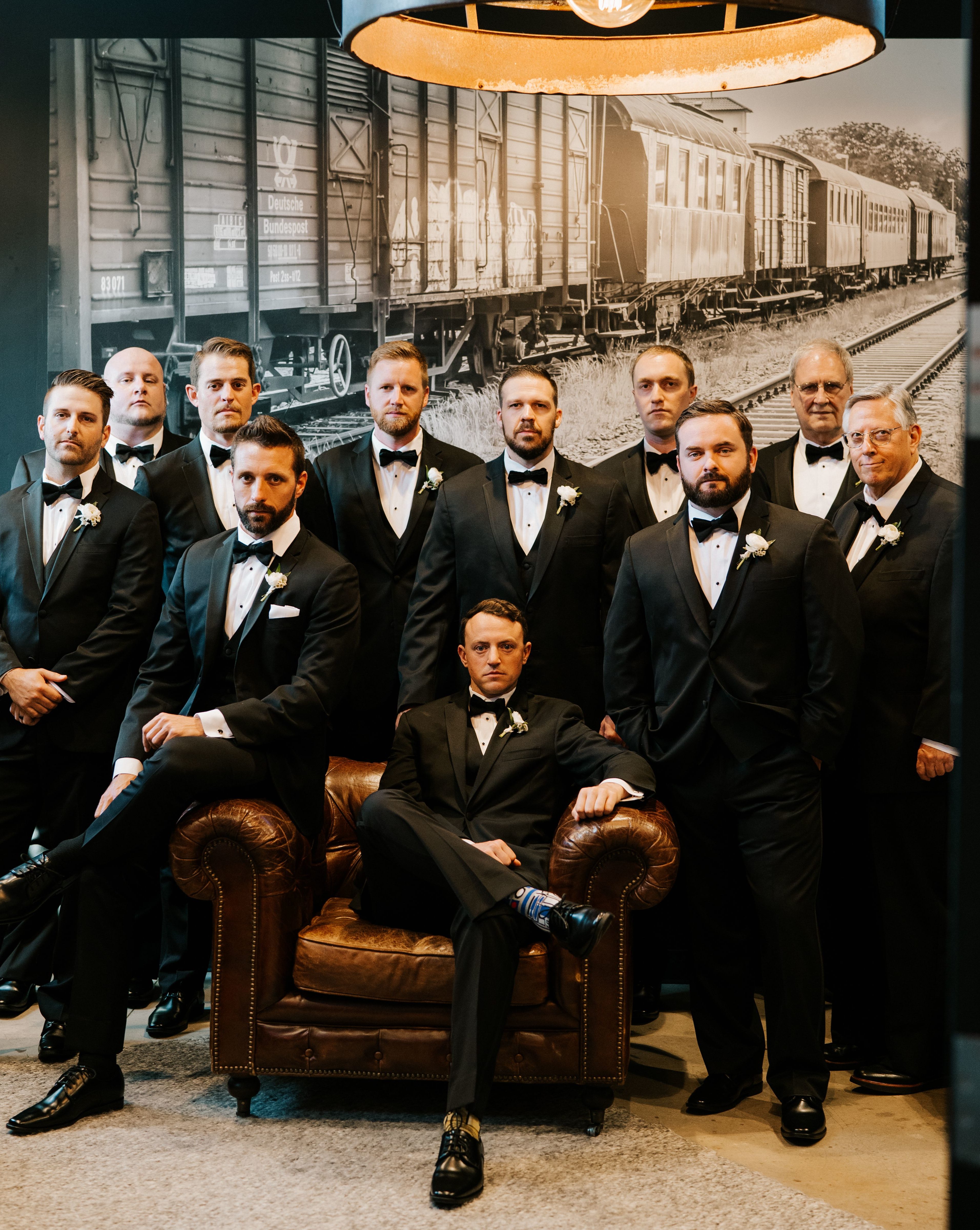 The groom sits in a leather armchair surrounded by his groomsmen, all in black Tuxedos.