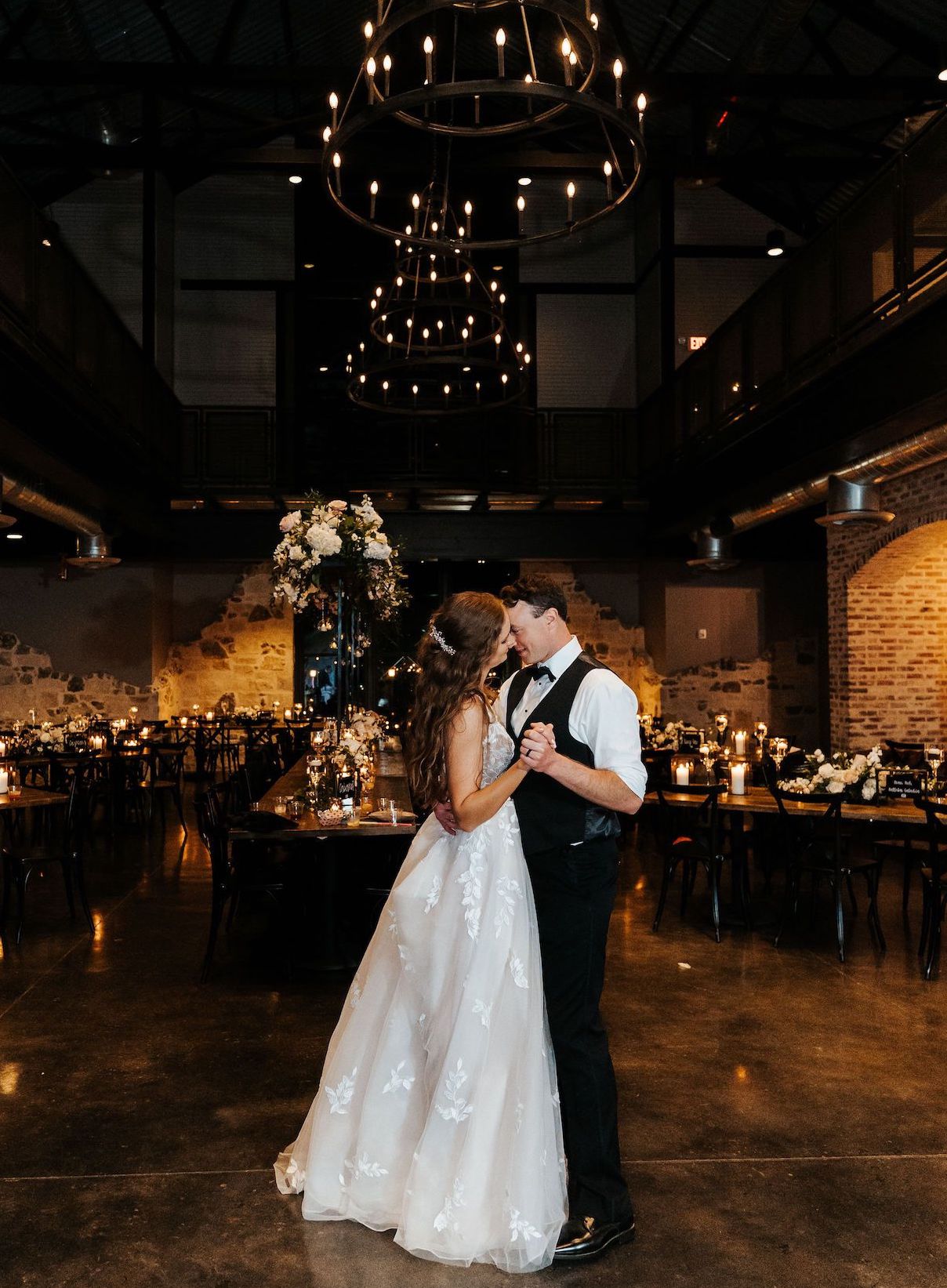 A beautiful couple shares a last dance in their empty reception venue, illuminated by candlelight and soft light from iron chandeliers.