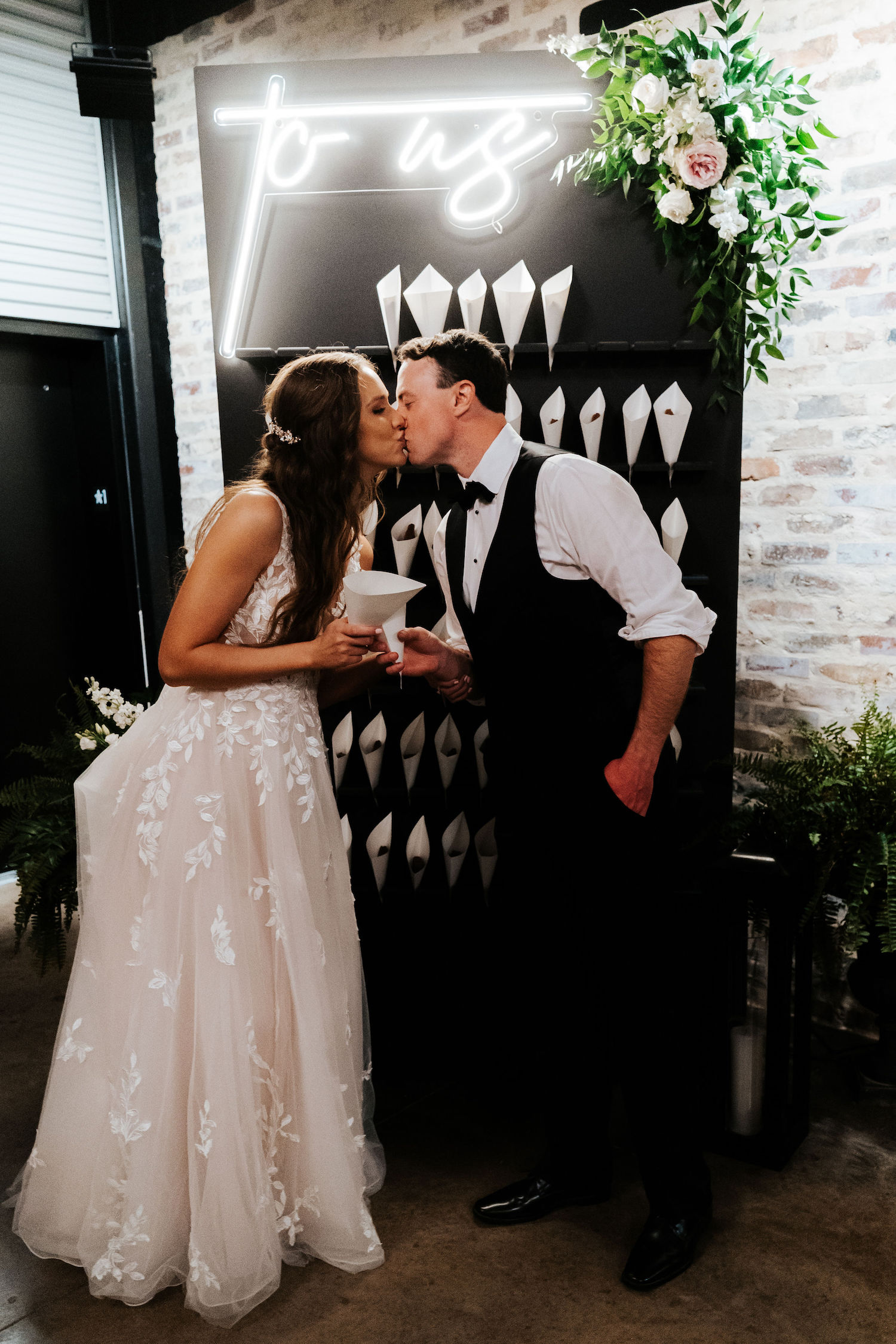 The newlywed couple exchanges a kiss in front of their custom brownie wall adorned with florals and a neon sign that reads "to us."