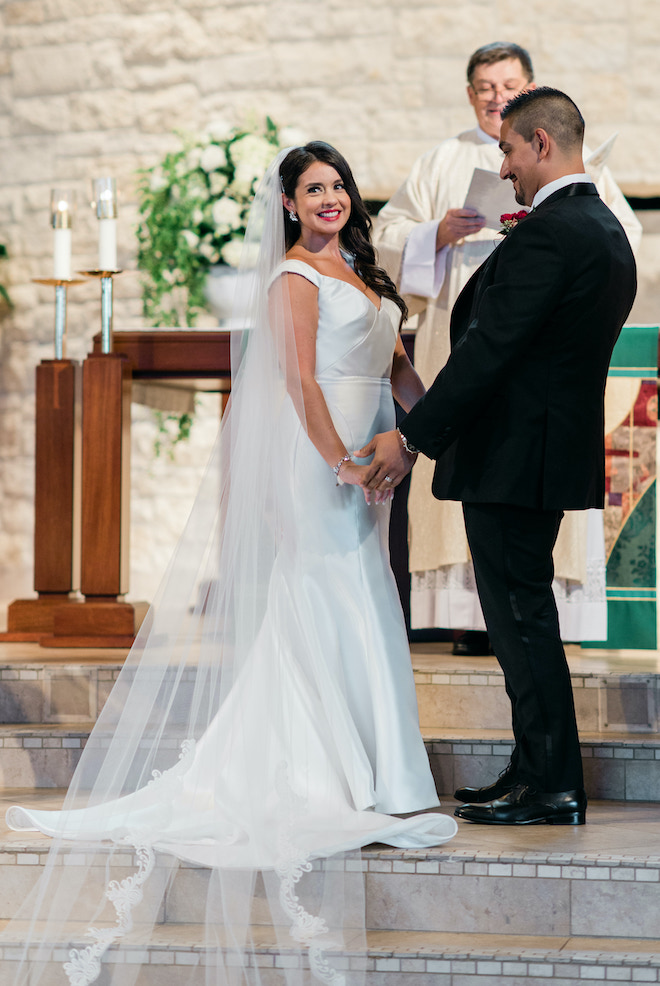 The bride smiles at the alter while the groom looks down at her.