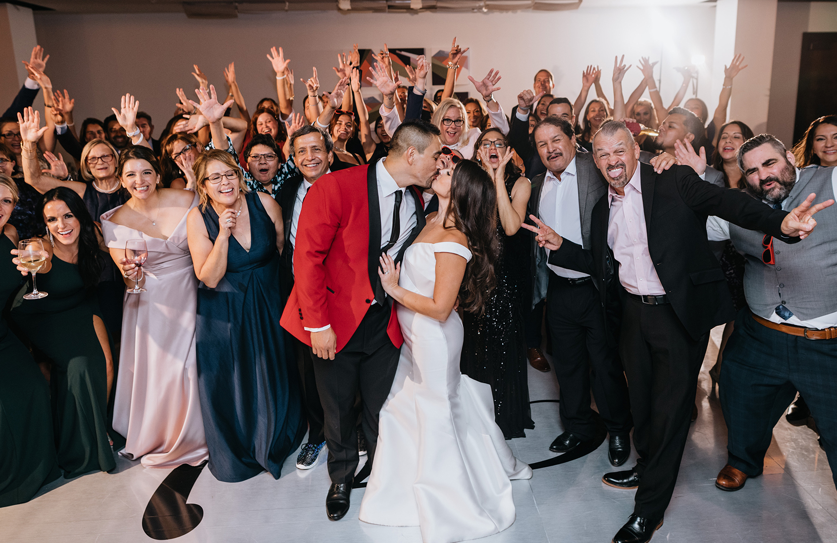 The bride and groom kiss on the dance floor with their wedding guests all behind them.