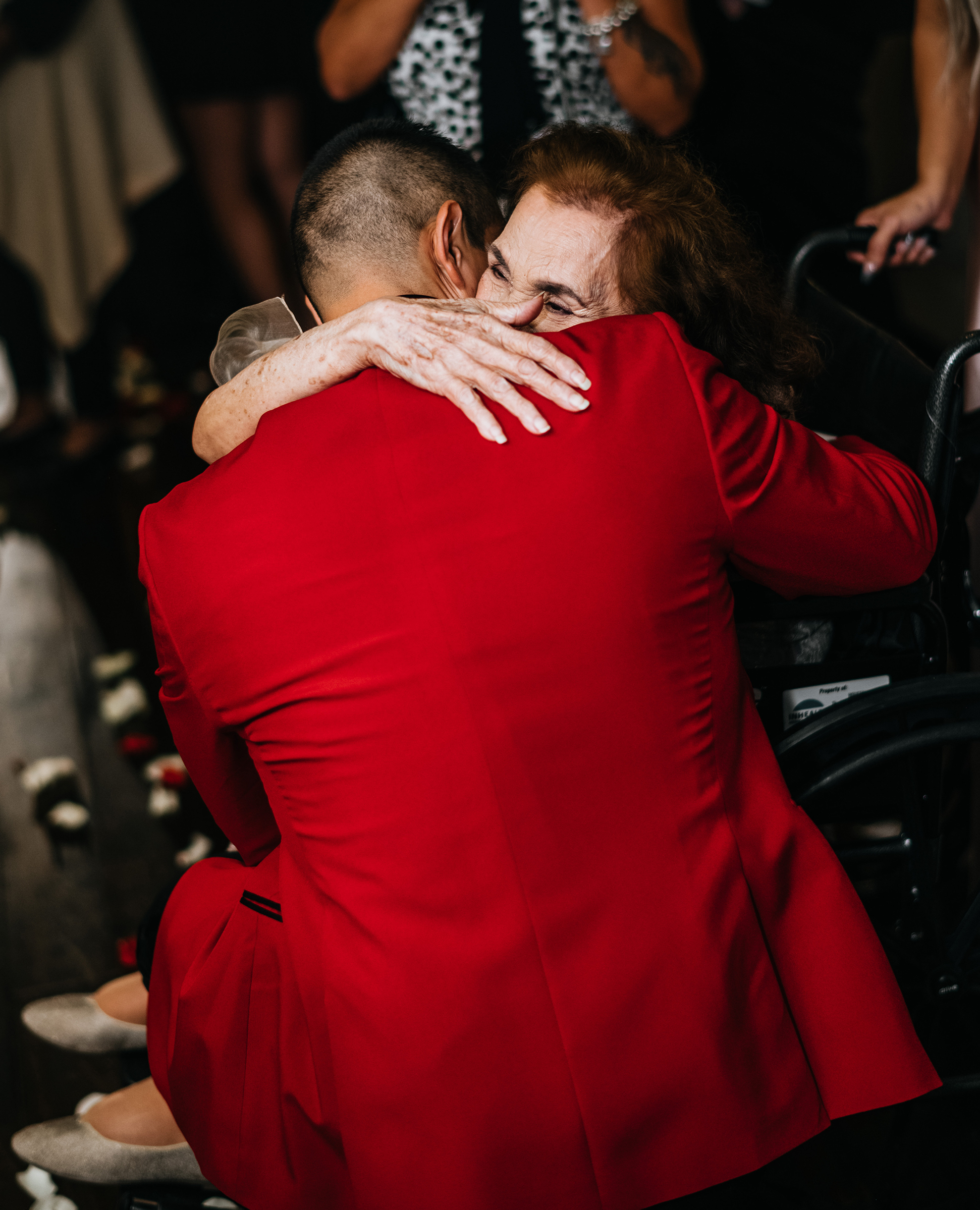 The groom is in a ruby red suitecoat hugging his grandmother who is in a wheelchair.