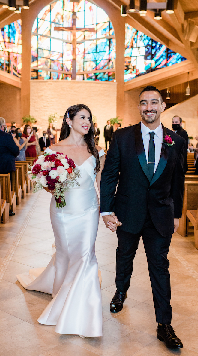 The bride, holding a ruby red, blush and ivory bouquet, holds hand with a groom in a black tuxedo after their wedding ceremony in Houston, Texas. 