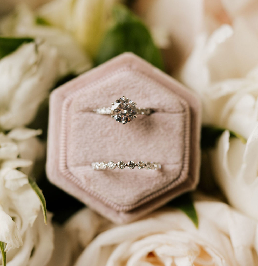 A bride's engagement ring and diamond wedding band rest in their box surrounded by white roses.