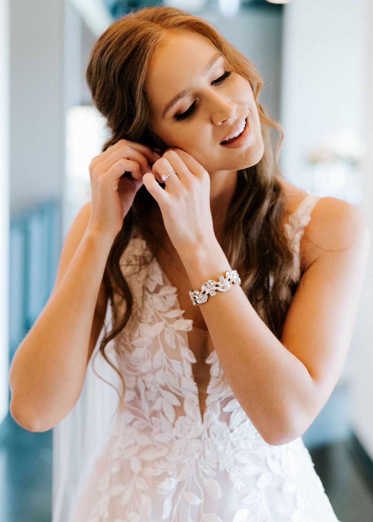 A bride in delicate, blush makeup and soft curls puts on her wedding earrings.