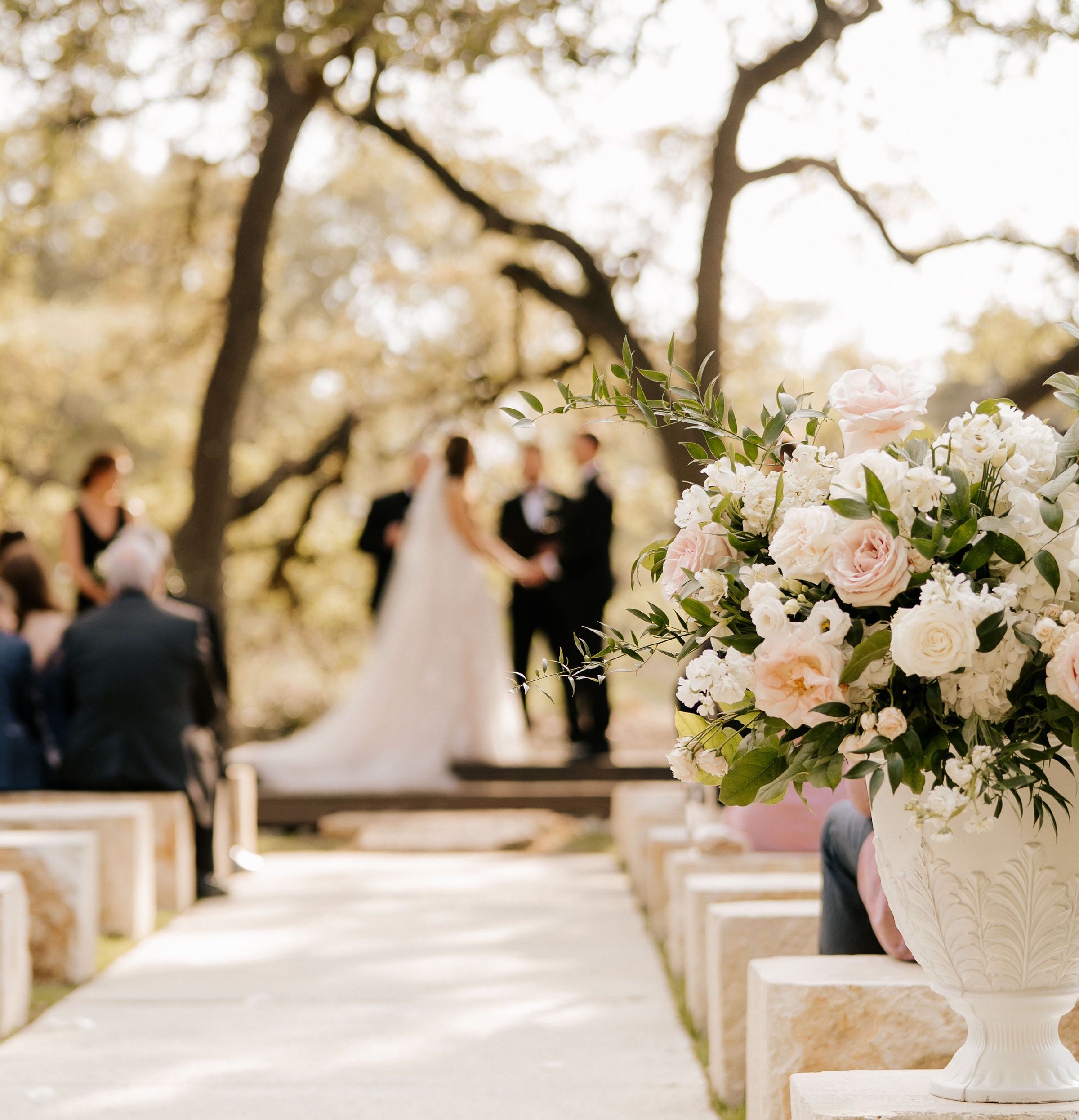 The couple stands under a canopy of trees in the distance while exchanging vows as a blush and white floral arrangement rests on a stone pew in focus.