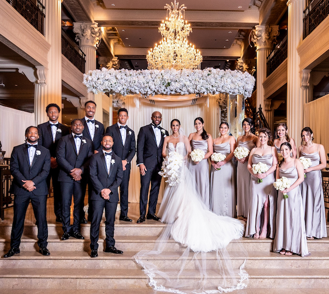 A photographical portrait of the bridesmaids, groomsmen, bride, and groom under a chandelier of the Corinthian Houston ballroom. 