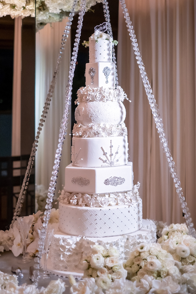 A white six tiered cake decorated in silver, crystal, and jewel like decorations and white roses. 