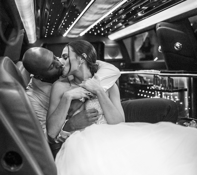 Groom and bride share a kiss as they embrace each other sitting inside of a limo. 