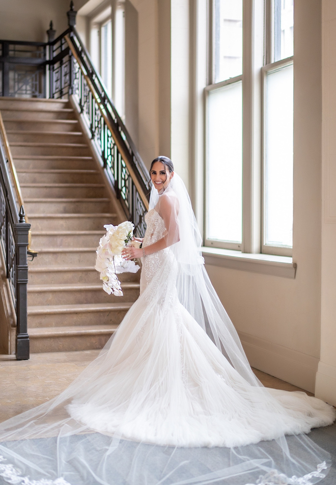 A bride is smiling as she holds her white floral bouquet and wears a long veil that extends across the floor.