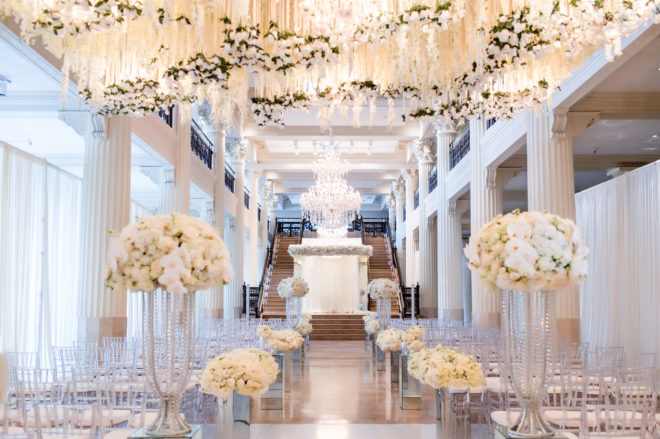 Ballroom of The Corinthian Houston decorated with many white florals hanging from the ceiling and down the aisle in floral arrangements. 