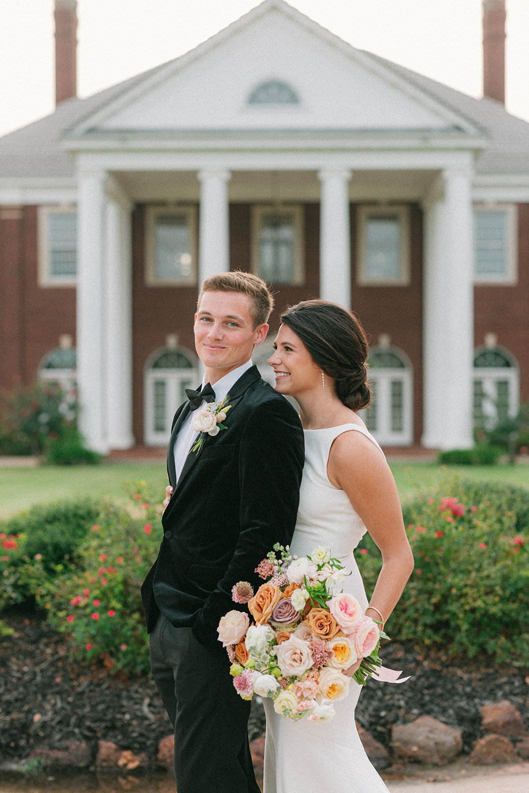 Bride smiles up at the groom while holding her artfully designed bridal bouquet. The couple is standing in front of the Mansion at ColoVista.