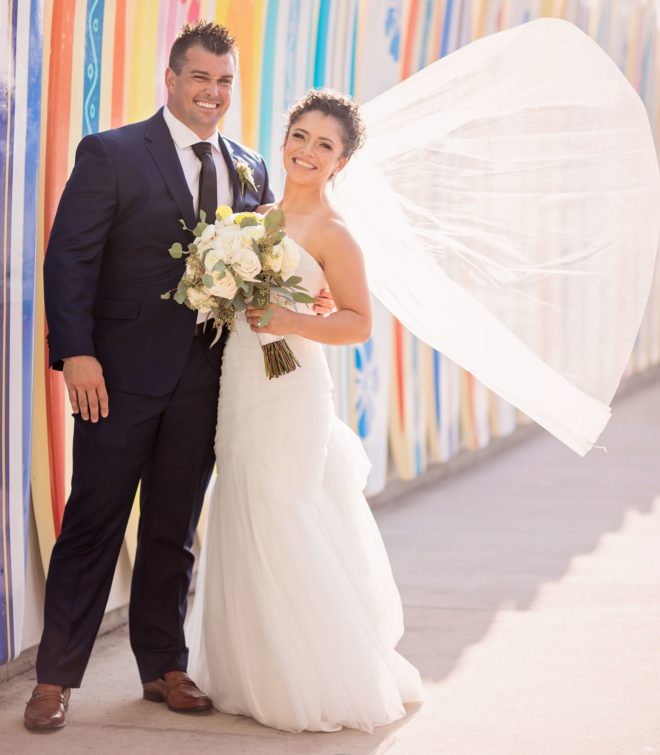 Groom and bride, with veil blowing in wind, pose in front of colorful outdoor surf boards at their lakeside wedding at Margaritaville Lake Resort, Lake Conroe and Houston. 