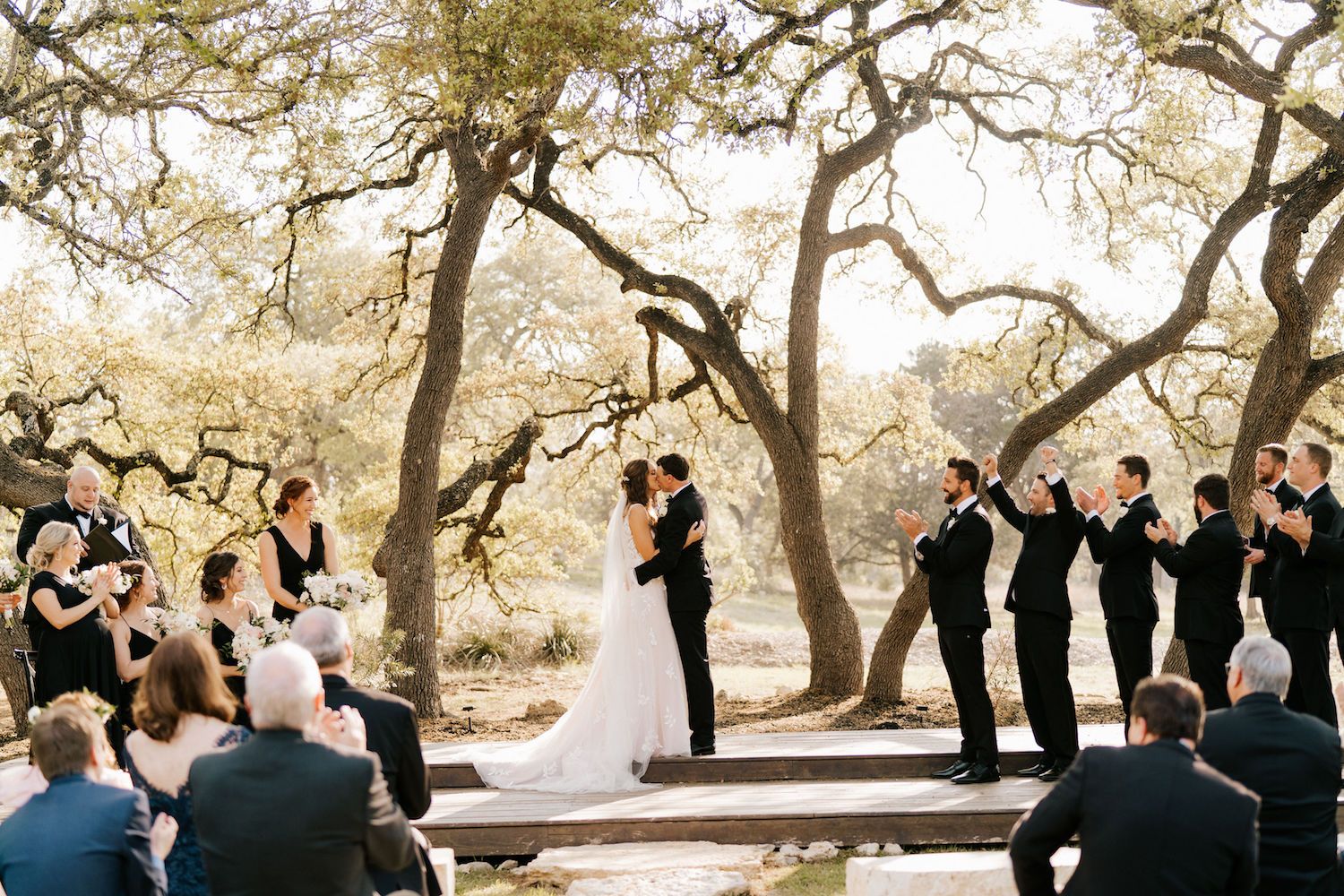 A newlywed couple exchanges a kiss under a natural arch of trees in Texas hill country.