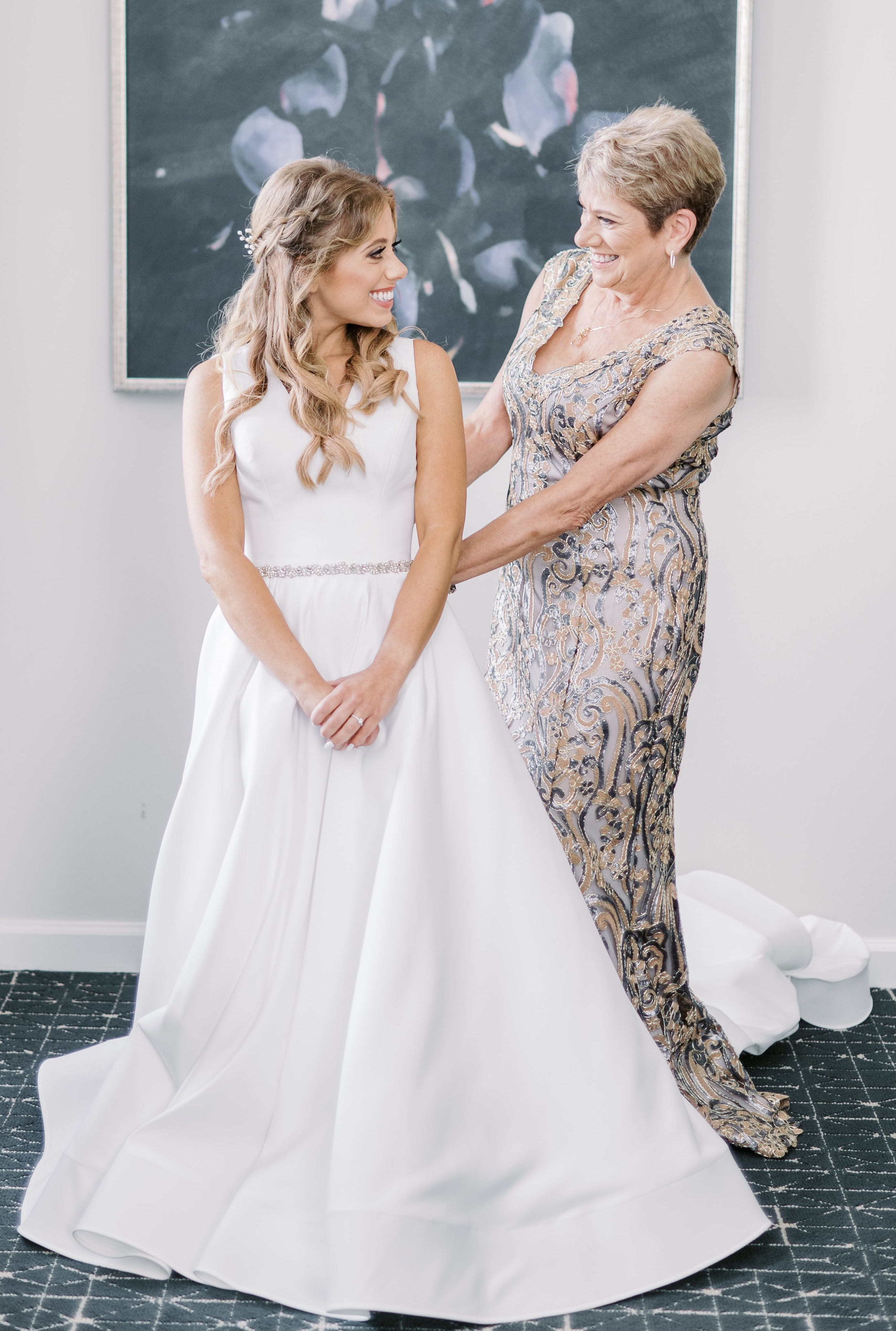 The bride smiles at her mom as she is putting on her white wedding gown.