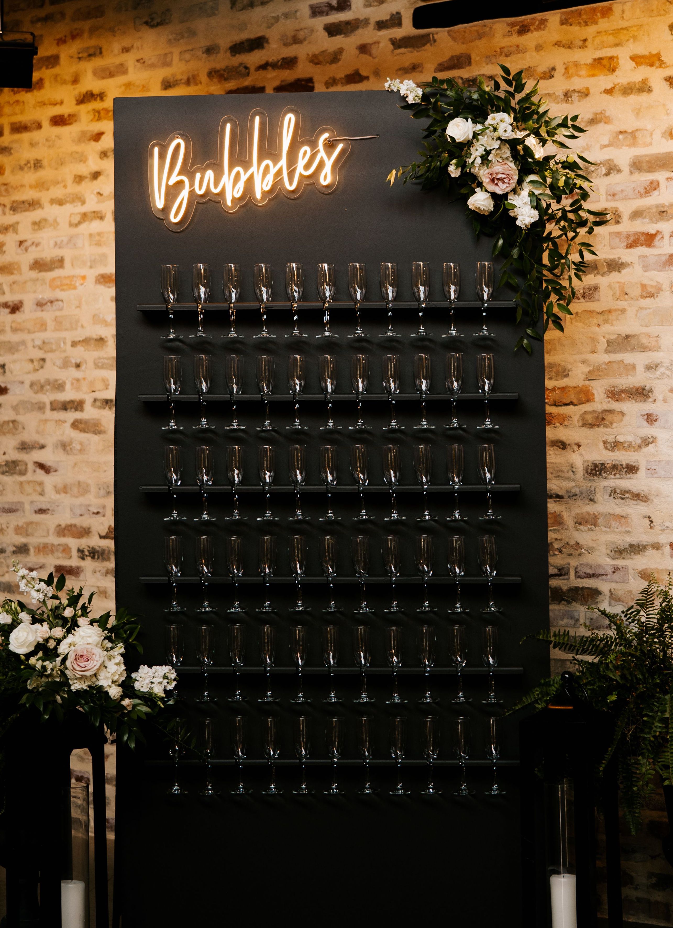 A black champagne wall installment is adorned with white and blush floral arrangements standing in front of a distressed brick wall.