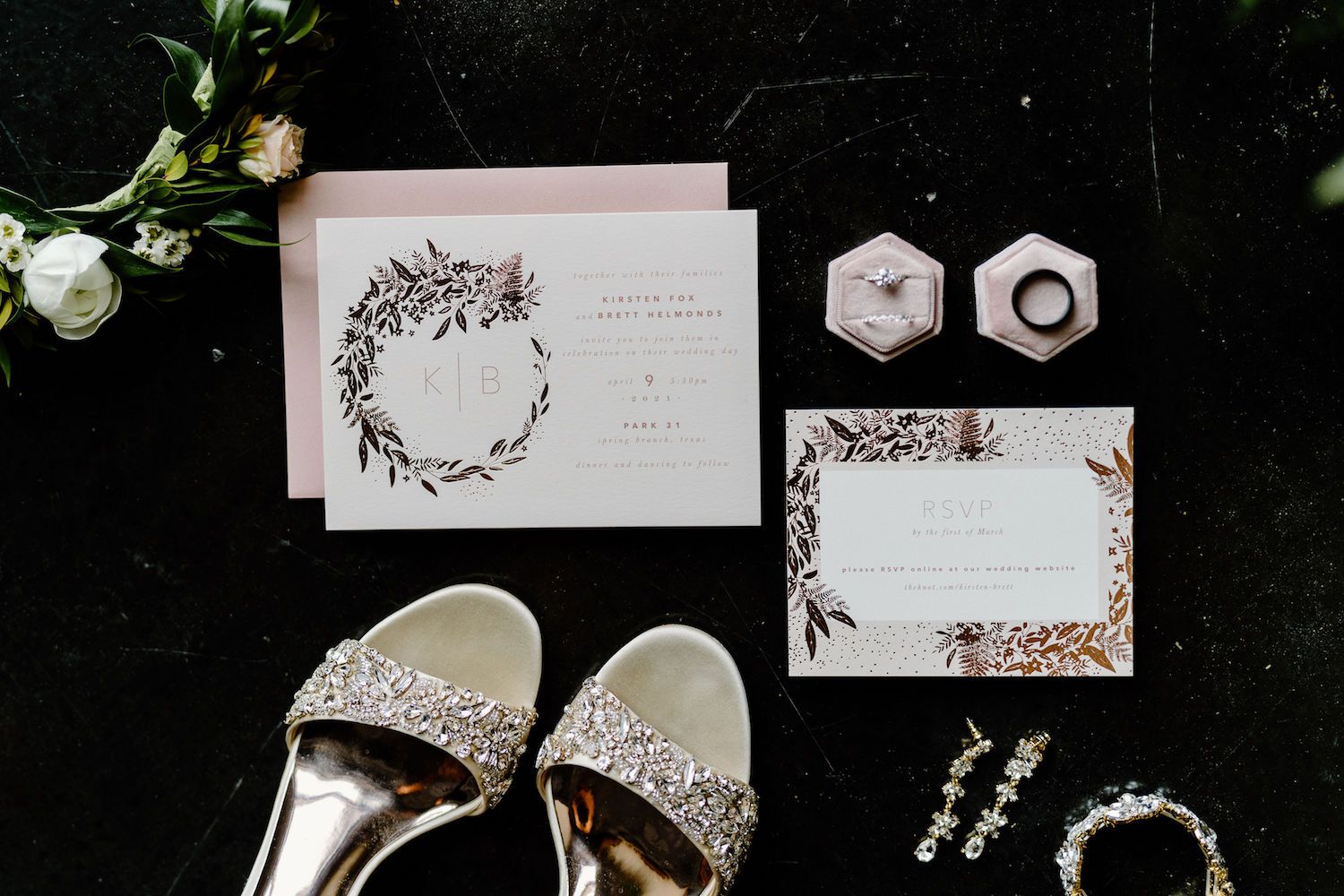 A flat lay showcases a couple's black, white, and blush floral wedding invitations, the bride's jewelry, and her dazzling wedding shoes.