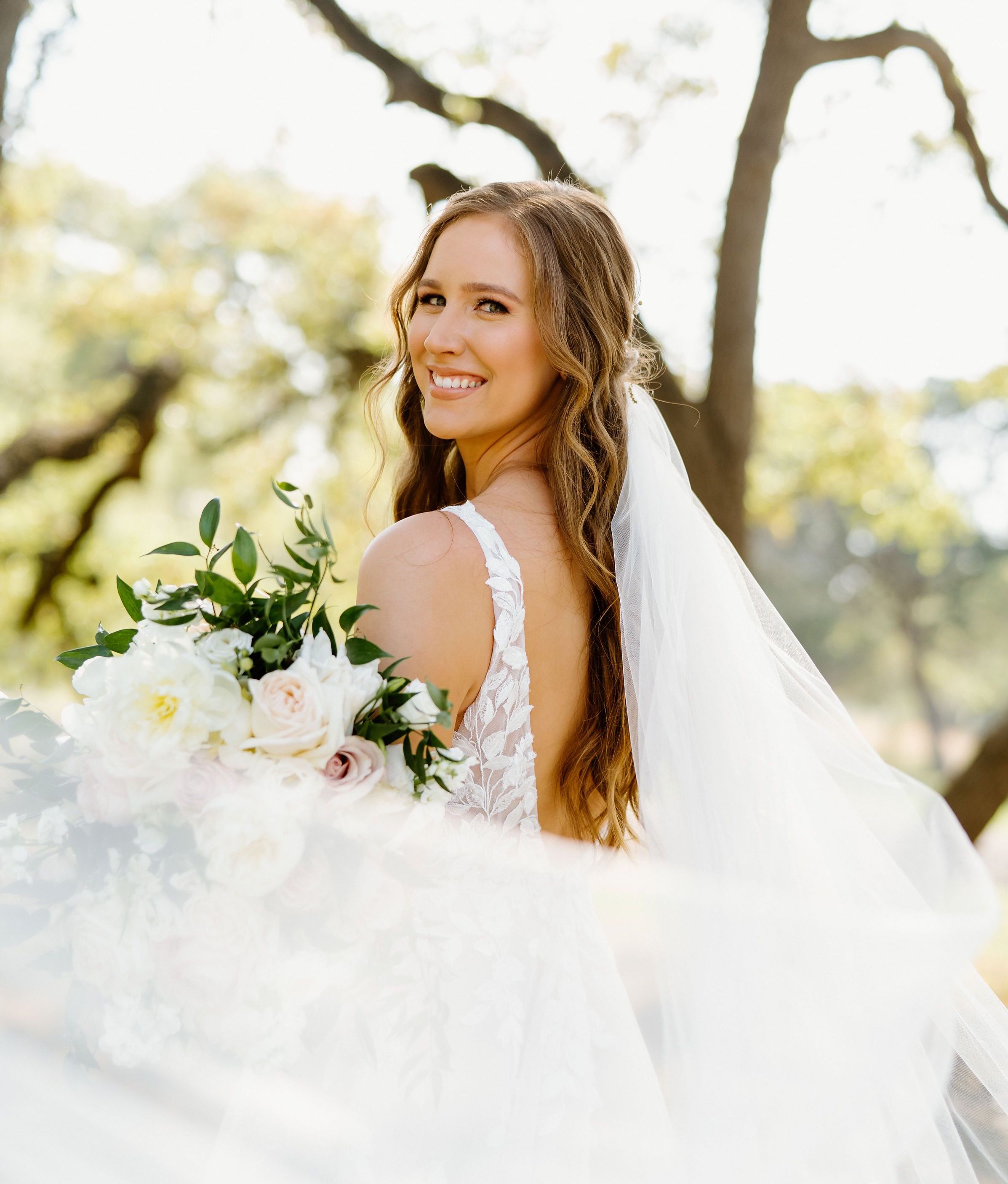 A beautiful bride is all smiles holding her bouquet as her cathedral length veil swirls out from behind her, catching the wind in a Texas Hill Country wedding designed by Scarlet Rose Events.