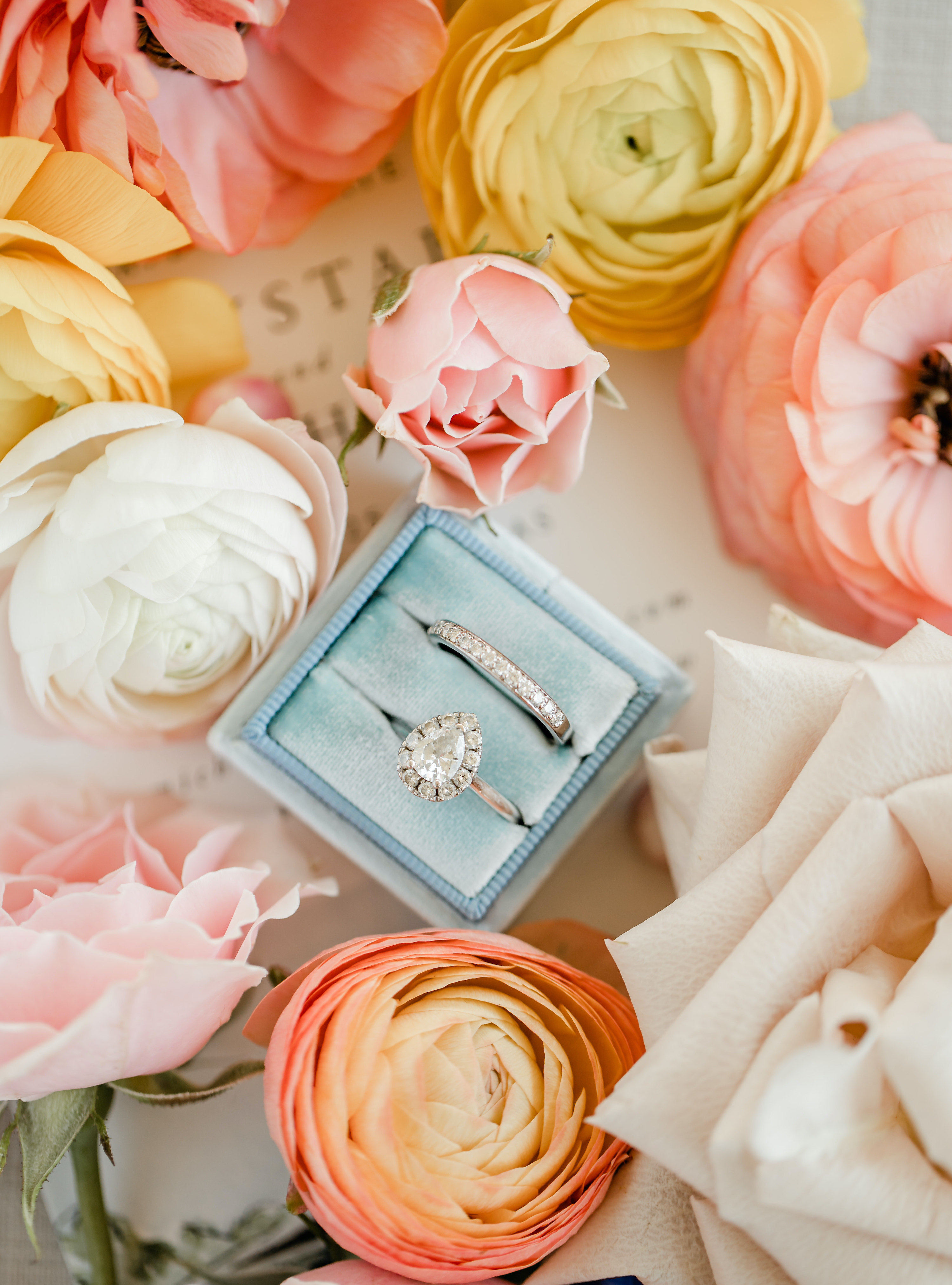 An engagement ring in a blue box surrounded by vibrant flowers.