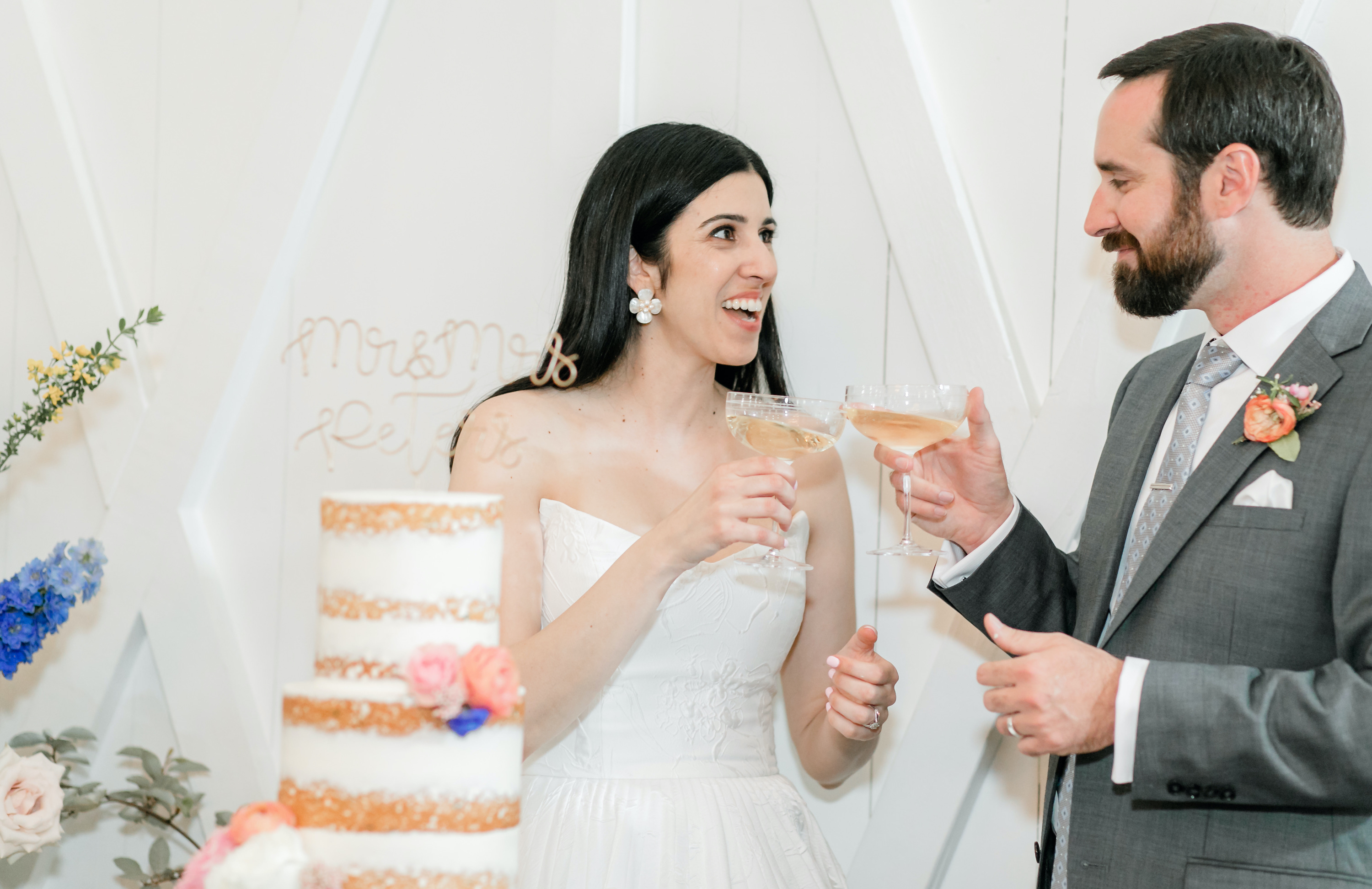 The bride smiles at the groom as they cheers glasses by their wedding cake.