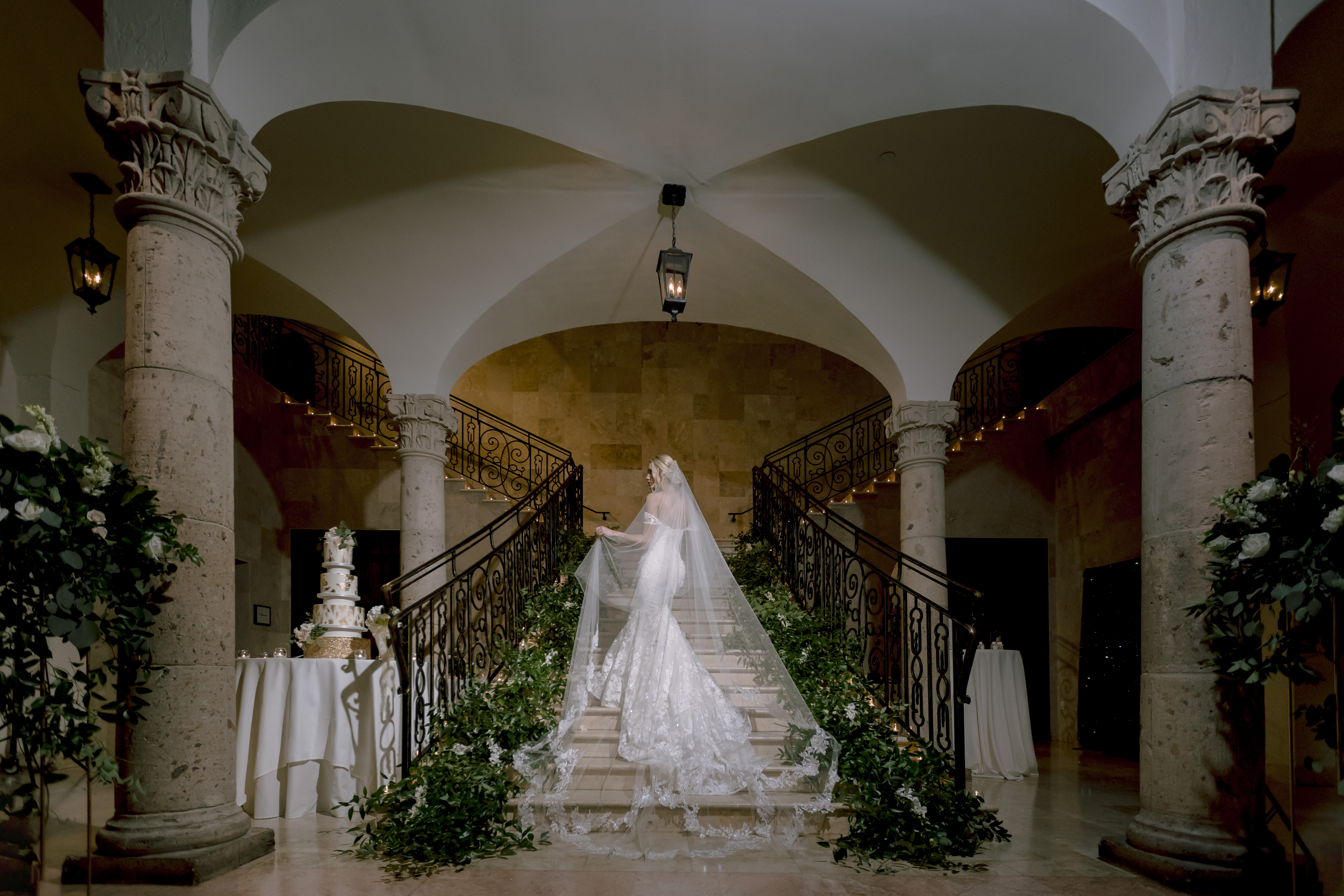 The bride poses in the middle of the grand staircase with her veil trailing behind her before her greenery-filled wedding.