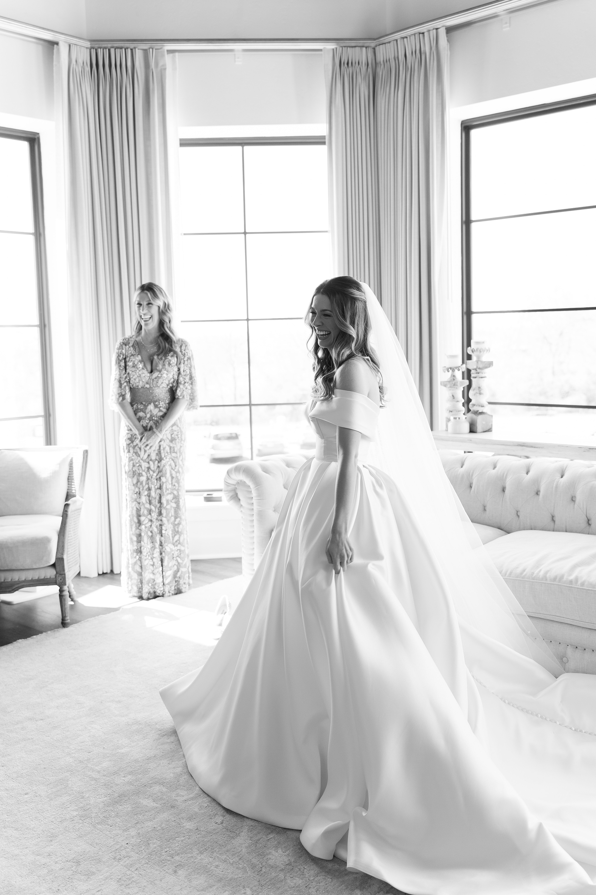 The bride smiles in her wedding dress while her mom is next to her in the bridal suite.